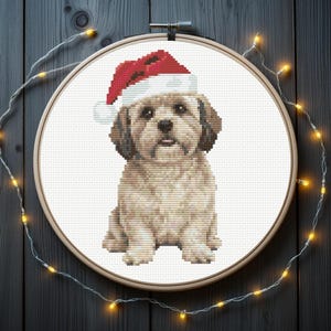 May include: A cross-stitch embroidery piece featuring a light brown dog wearing a red and white Santa hat, displayed within a circular wooden embroidery hoop. The design is centred on a white fabric background, showcasing detailed needlework.