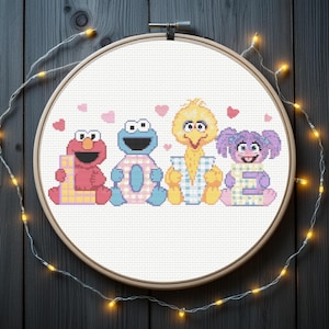 May include: A cross-stitch hoop art featuring Elmo, Cookie Monster, Big Bird, and Abby Cadabby. Each character holds a letter, with pink hearts above. The characters are in a row, with a wooden hoop and string lights.