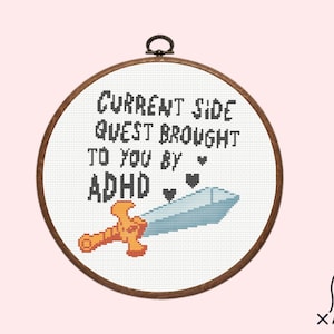 May include: A white embroidery hoop with a brown wooden frame. The hoop contains a cross-stitch design that reads "Current Side Quest Brought To You By ADHD" with a small sword and hearts.