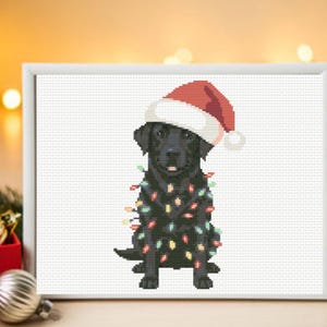 May include: A cross-stitch pattern featuring a black Labrador retriever wearing a red and white Santa hat, wrapped in a string of multicolored Christmas lights, displayed in a white rectangular frame against a blurred holiday background.