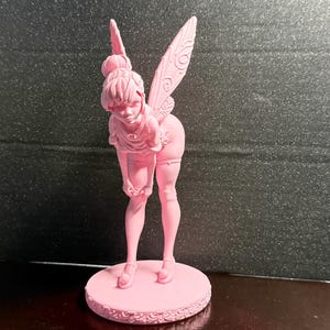 May include: A pink figurine of a fairy with detailed wings, a bun hairstyle, and a playful pose. The fairy is standing on a round base with a decorative edge. The figurine is made of a solid material.