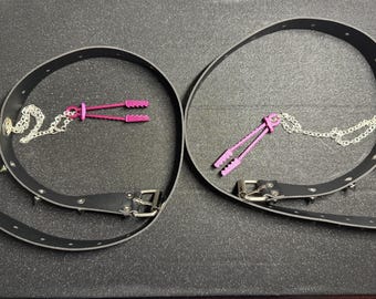May include: Two black leather chokers with silver hardware and studded accents. Each choker features a silver chain and a pink clamp-style accessory. The items are displayed on a dark gray surface.