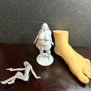 May include: The image showcases three distinct figurines: a light gray figure of a woman, a silver figure of a woman, and a tan foot. The woman figure is standing on a white base. The silver figure is lying down. The foot is a solid color.