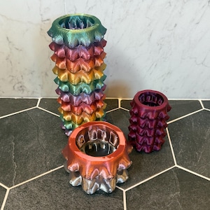 May include: Three decorative, colorful, 3D-printed vases. The tallest vase is a rainbow of colors, while the other two are smaller and in shades of red and purple. The vases have a textured, spiky design.
