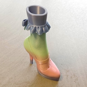 May include: A whimsical, 3D-printed shoe-shaped vase. The design features a high heel, a pink and peach shoe, and a green and yellow fishnet stocking. A gray cylinder with a blue ruffled trim forms the top of the vase.