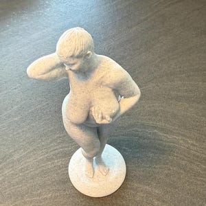 Peut inclure: Une figurine blanc cassé mouchetée représentant une personne debout, une main derrière la tête et l'autre tendue vers l'avant. La sculpture présente des formes arrondies et généreuses, fixée sur une base circulaire avec une finition texturée.