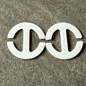 May include: Two white, semi-circular bra strap holders with a vertical bar in the centre. The bra strap holders are made of a felt-like material and are positioned side-by-side on a grey surface.