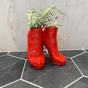 May include: A pair of vibrant red high-heeled shoe planters, each holding green succulents. The glossy finish of the shoes contrasts with the natural greenery, creating a striking decorative accent. The planters are set on a black and white tiled surface.