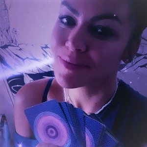 May include: A woman holds a deck of blue and purple tarot cards with a swirling, eye-like design. The cards are arranged in a fan shape, revealing the backs of the cards.