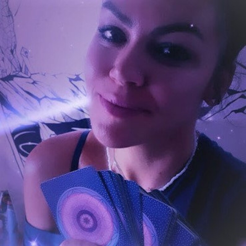 May include: A woman holds a deck of blue and purple tarot cards with a swirling, eye-like design. The cards are arranged in a fan shape, revealing the backs of the cards.