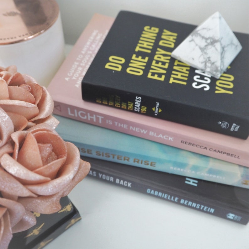 May include: A stack of four books with pink, blue, and black covers. The top book is black with yellow text that reads "Do One Thing Every Day That Scares You." The second book is pink with white text that reads "Light is the New Black." The third book is blue with white text that reads "Rise Sister Rise." The bottom book is black with white text that reads "Has Your Back."
