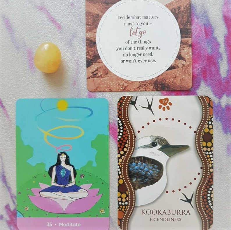 May include: Three cards from a deck of oracle cards. The first card is a light brown card with a white circle containing the text "Decide what matters most to you - let go of the things you don't really want, no longer need, or won't ever use.". The second card is a blue and green card with a woman meditating in a lotus position. The text on the card reads "35. Meditate". The third card is a brown card with a white bird with blue feathers and the text "19 Kookaburra Friendliness".
