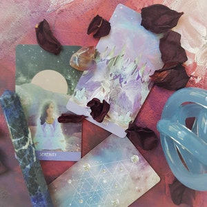 May include: Three tarot cards on a pink and red background with dried rose petals. The cards are "The Magician", "Serenity", and a card with a geometric pattern. A blue glass orb is in the bottom right corner.