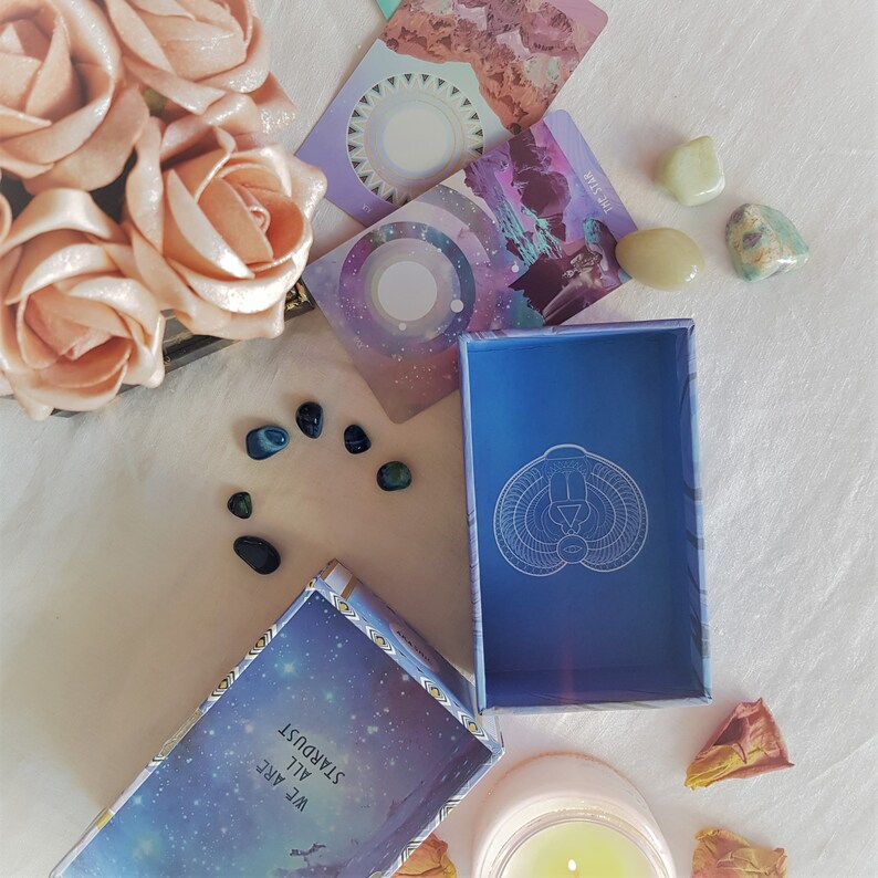 May include: A blue box with a white design of a stylized figure with a triangle inside a circle. The box is open and sitting on a white surface. There are several small blue and green stones scattered around the box. There are also two cards with a purple and pink background and a white circle in the center. The text on the cards reads "The Stars" and "We Are All Stardust".