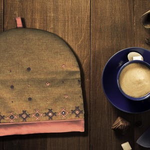May include: A brown tea cozy with pink trim and embroidered details sits on a wooden surface next to a blue saucer with a cup of coffee.