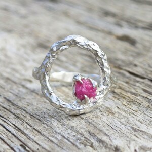 May include: A silver ring with a textured, organic design. The ring features a bright pink, raw gemstone set within the circular frame. The ring is displayed on a wooden surface.