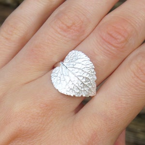 May include: A silver ring shaped like a leaf with detailed veins. The ring is worn on a person's finger.