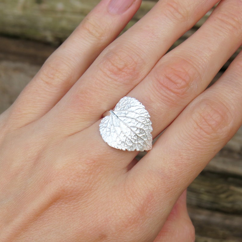 May include: A silver ring shaped like a leaf with detailed veins. The ring is worn on a person's finger.