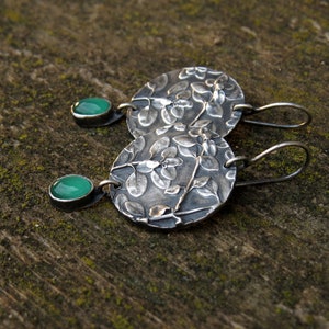 Puede incluir: Un par de pendientes de plata con un dise&ntilde;o floral y detalles de piedras preciosas verdes.