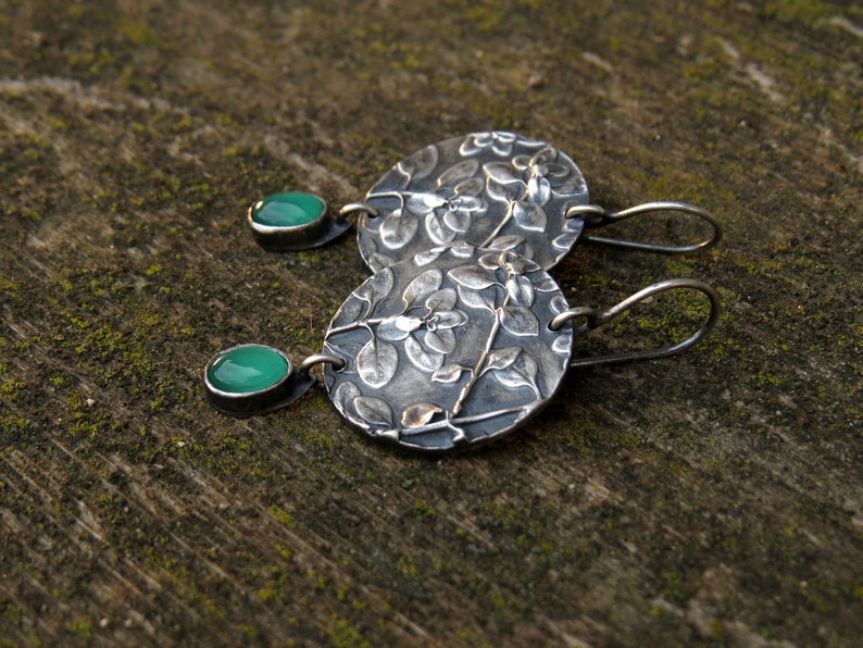 Puede incluir: Un par de pendientes de plata con un dise&ntilde;o floral y detalles de piedras preciosas verdes.