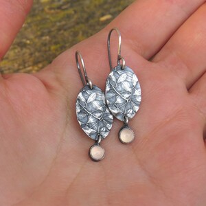 May include: A pair of silver-toned dangle earrings. Each earring features an oval-shaped, textured silver pendant with a leaf design, and a small, round, pale pink stone. The earrings are displayed against a blurred background.