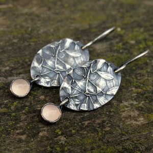 May include: A pair of silver earrings with an oval, leaf-patterned design. Each earring features a small, round, pale pink stone. The earrings are displayed on a textured, mossy surface.