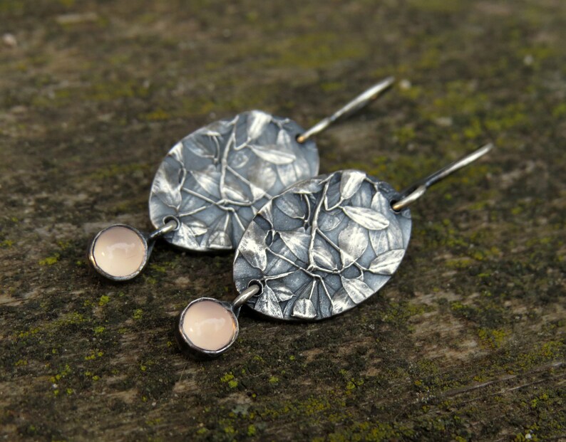 May include: A pair of silver earrings with an oval, leaf-patterned design. Each earring features a small, round, pale pink stone. The earrings are displayed on a textured, mossy surface.