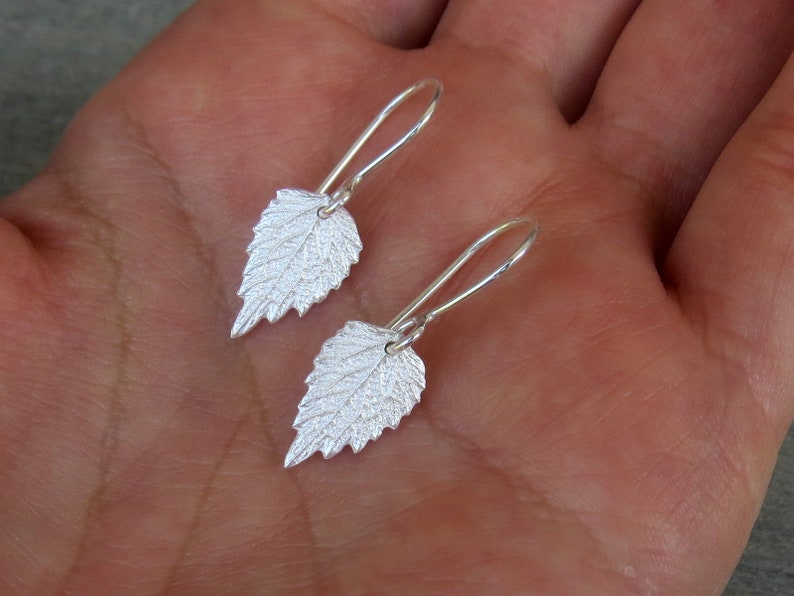 May include: A pair of silver earrings shaped like leaves. The leaves have a textured surface and are hanging from silver hooks.