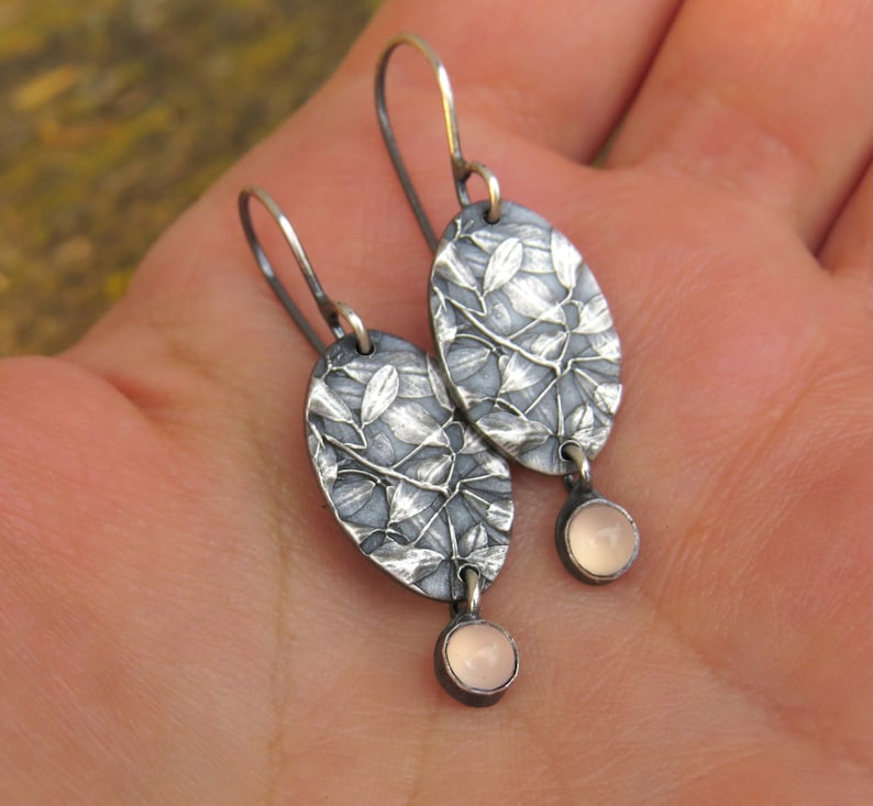 May include: Silver oval earrings with a leaf design and small, round, pink gemstones. The earrings have a textured surface and a hook closure. The gemstones are a soft pink color.