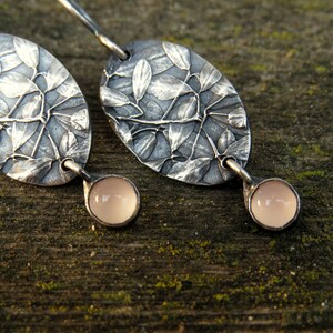 May include: A pair of silver earrings with an oval leaf design and a small, round, pale pink stone at the bottom. The earrings are set against a textured wooden surface.