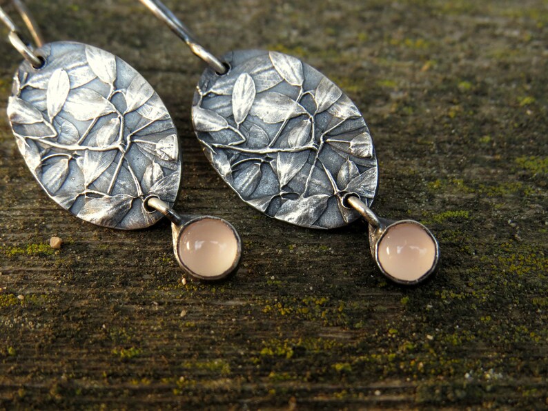 May include: A pair of silver earrings with an oval leaf design and a small, round, pale pink stone at the bottom. The earrings are set against a textured wooden surface.