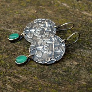 Puede incluir: Un par de pendientes de plata con un dise&ntilde;o floral y detalles de piedras preciosas verdes.