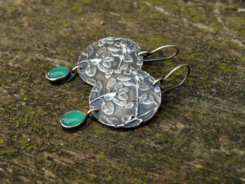 Puede incluir: Un par de pendientes de plata con un dise&ntilde;o floral y detalles de piedras preciosas verdes.