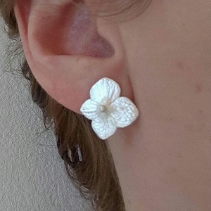 Könnte beinhalten: Ein Paar weiße Ohrringe in Blütenform. Die Ohrringe sind aus einem strukturierten Material gefertigt und haben ein zartes, feminines Design.