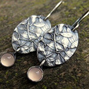 May include: A pair of silver-toned earrings. Each earring features an oval-shaped design with a leaf pattern and a small, round, light pink stone at the bottom. The earrings are displayed on a textured, mossy surface.