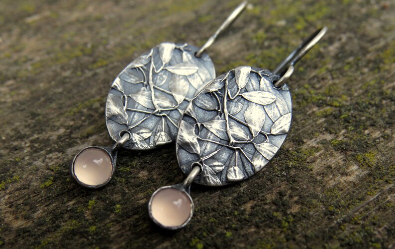 May include: A pair of silver-toned earrings. Each earring features an oval-shaped design with a leaf pattern and a small, round, light pink stone at the bottom. The earrings are displayed on a textured, mossy surface.
