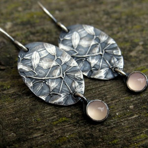 May include: A pair of silver earrings with an oval leaf design. The earrings have a dark gray background with a detailed leaf pattern. Each earring has a small, round, light pink stone dangling from the bottom.