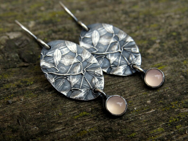May include: A pair of silver earrings with an oval leaf design. The earrings have a dark gray background with a detailed leaf pattern. Each earring has a small, round, light pink stone dangling from the bottom.
