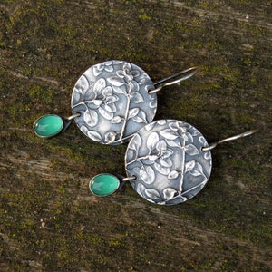 Puede incluir: Un par de pendientes de plata con un dise&ntilde;o floral y detalles de piedras preciosas verdes.