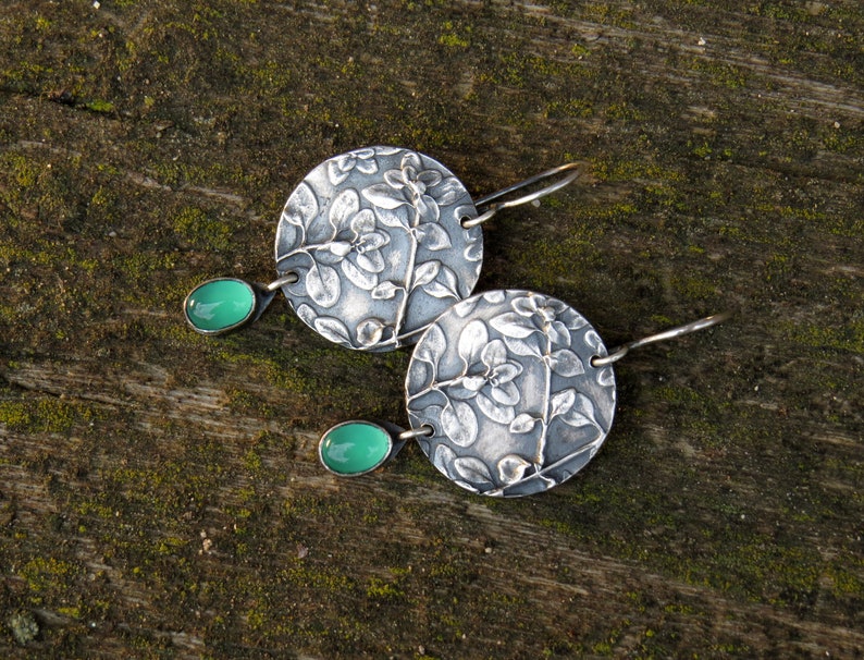 Puede incluir: Un par de pendientes de plata con un dise&ntilde;o floral y detalles de piedras preciosas verdes.