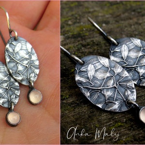 May include: A pair of silver earrings with an oval leaf design and a small, round, light pink stone at the bottom. The earrings are on a silver hook. The design is detailed and textured.