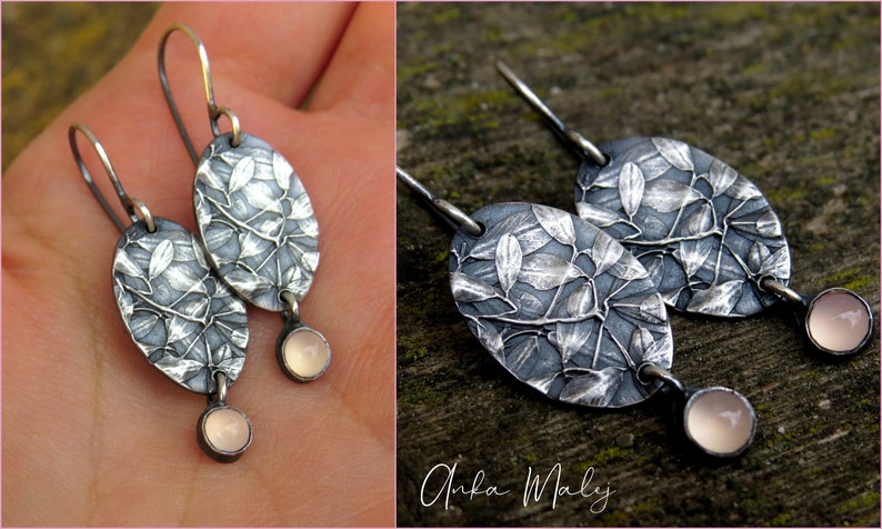 May include: A pair of silver earrings with an oval leaf design and a small, round, light pink stone at the bottom. The earrings are on a silver hook. The design is detailed and textured.