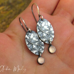 May include: Silver-toned oval earrings with a leaf design and small, round, pink gemstones. The earrings have a textured surface and a hook closure. The earrings are approximately 1.5 inches long.
