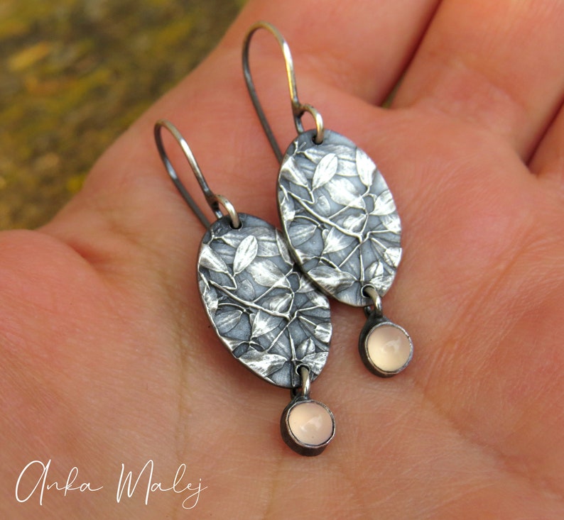 May include: Silver-toned oval earrings with a leaf design and small, round, pink gemstones. The earrings have a textured surface and a hook closure. The earrings are approximately 1.5 inches long.