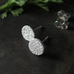 May include: A pair of silver stud earrings with a textured, circular design. The earrings are small and delicate.