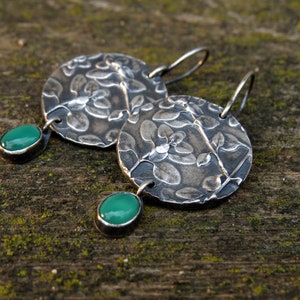 Puede incluir: Un par de pendientes de plata con un dise&ntilde;o floral y detalles de piedras preciosas verdes.