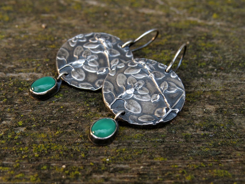 Puede incluir: Un par de pendientes de plata con un dise&ntilde;o floral y detalles de piedras preciosas verdes.