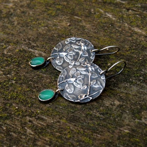 Puede incluir: Un par de pendientes de plata con un dise&ntilde;o floral y detalles de piedras preciosas verdes.