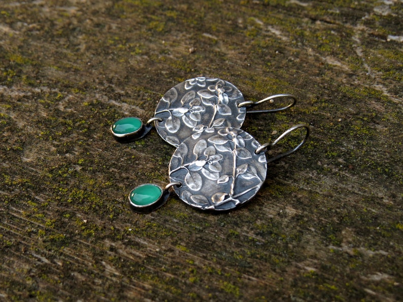Puede incluir: Un par de pendientes de plata con un dise&ntilde;o floral y detalles de piedras preciosas verdes.