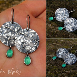 Puede incluir: Pendientes de plata con dise&ntilde;o floral y gotas de piedras preciosas verdes. Los pendientes se muestran en una mano y sobre una superficie de madera.